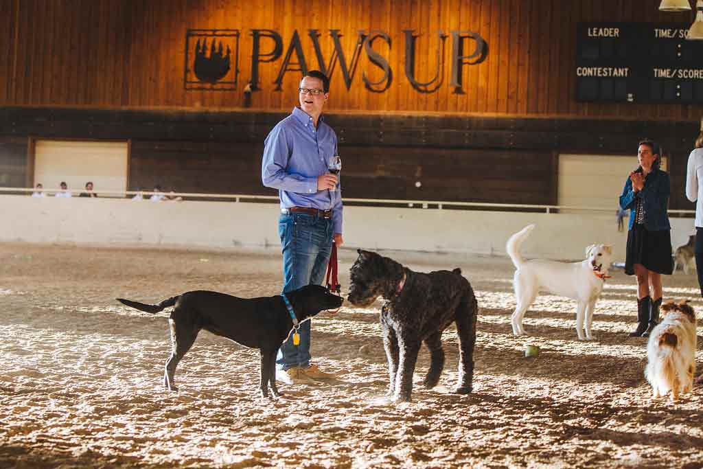 A man standing in a corral with several dogs playing around him. A sign on the wall behind them reads "Paws Up"