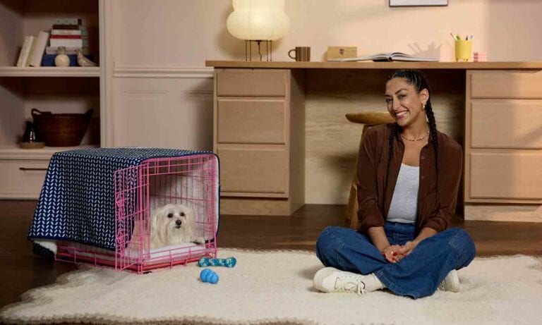 A woman sitting on the floor beside her dog, who is laying in a dog crate